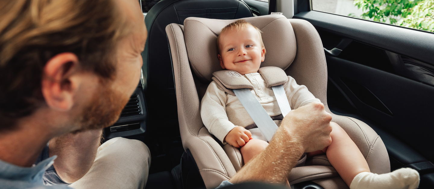Hymyilev&auml; vauva istuu turvallisesti teev&auml;risess&auml; Britax R&ouml;mer Dualfix 5Z -turvaistuimessa, joka on asennettu auton takapenkille. Sivulta n&auml;kyv&auml; aikuinen pit&auml;&auml; hell&auml;sti vauvan jalkoja kiinni samalla kun leikkii h&auml;nen kanssaan.